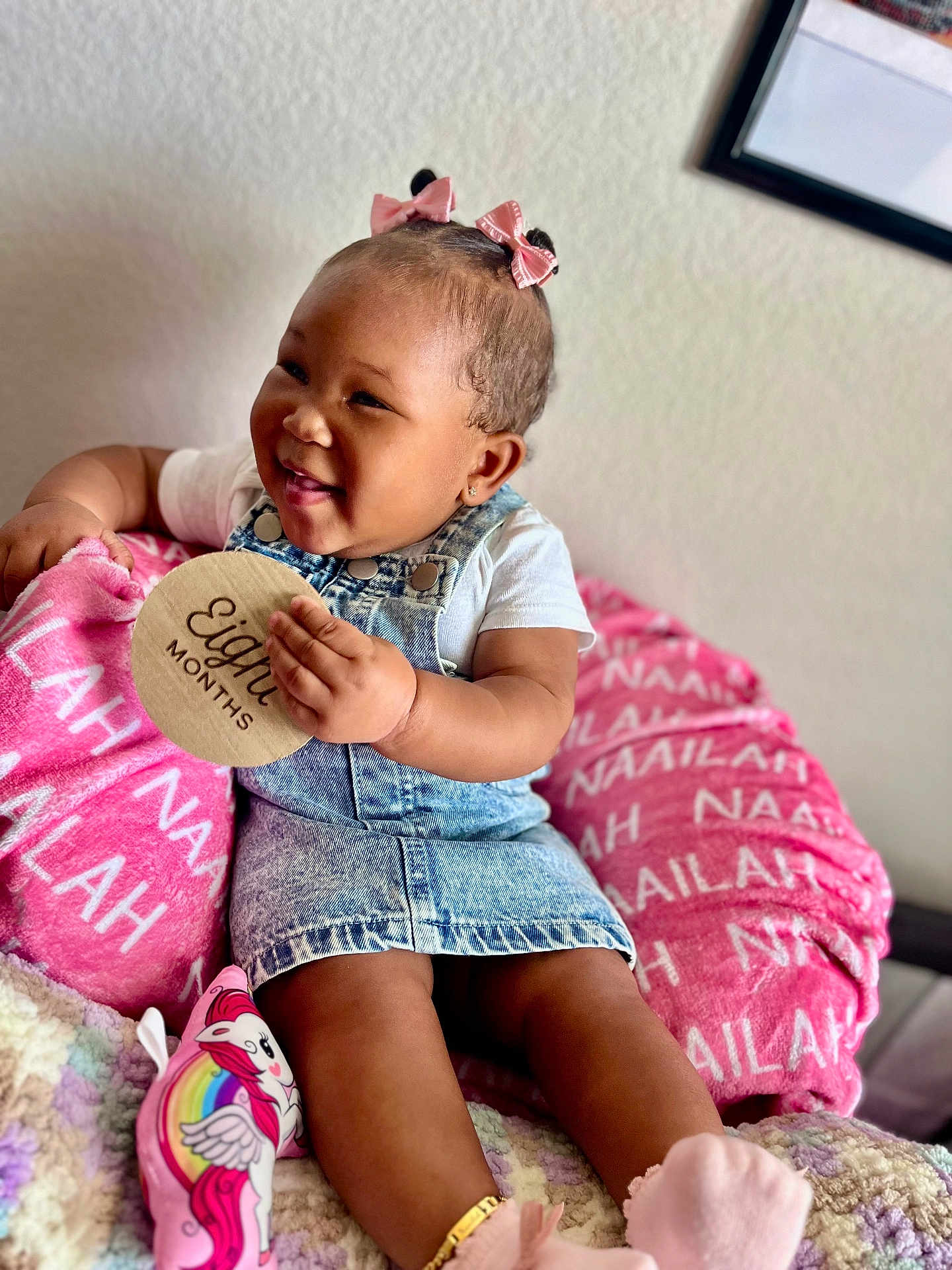 Naailah joined the competition — help win amazing prizes! baby, child, girl, smiling, denim_dress, pink_bows, cushion, personalized_blanket, toy, unicorn, wooden_sign, eight_months, sitting, happy, indoors, bracelet, white_shirt, cozy, portrait, cute