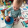 baby, blonde_hair, child, clothing, colorful, container, face, floor, floor_mat, formula, hand, head, heart_pattern, indoor, lid, long_sleeve_shirt, pants, playing, toddler, toy