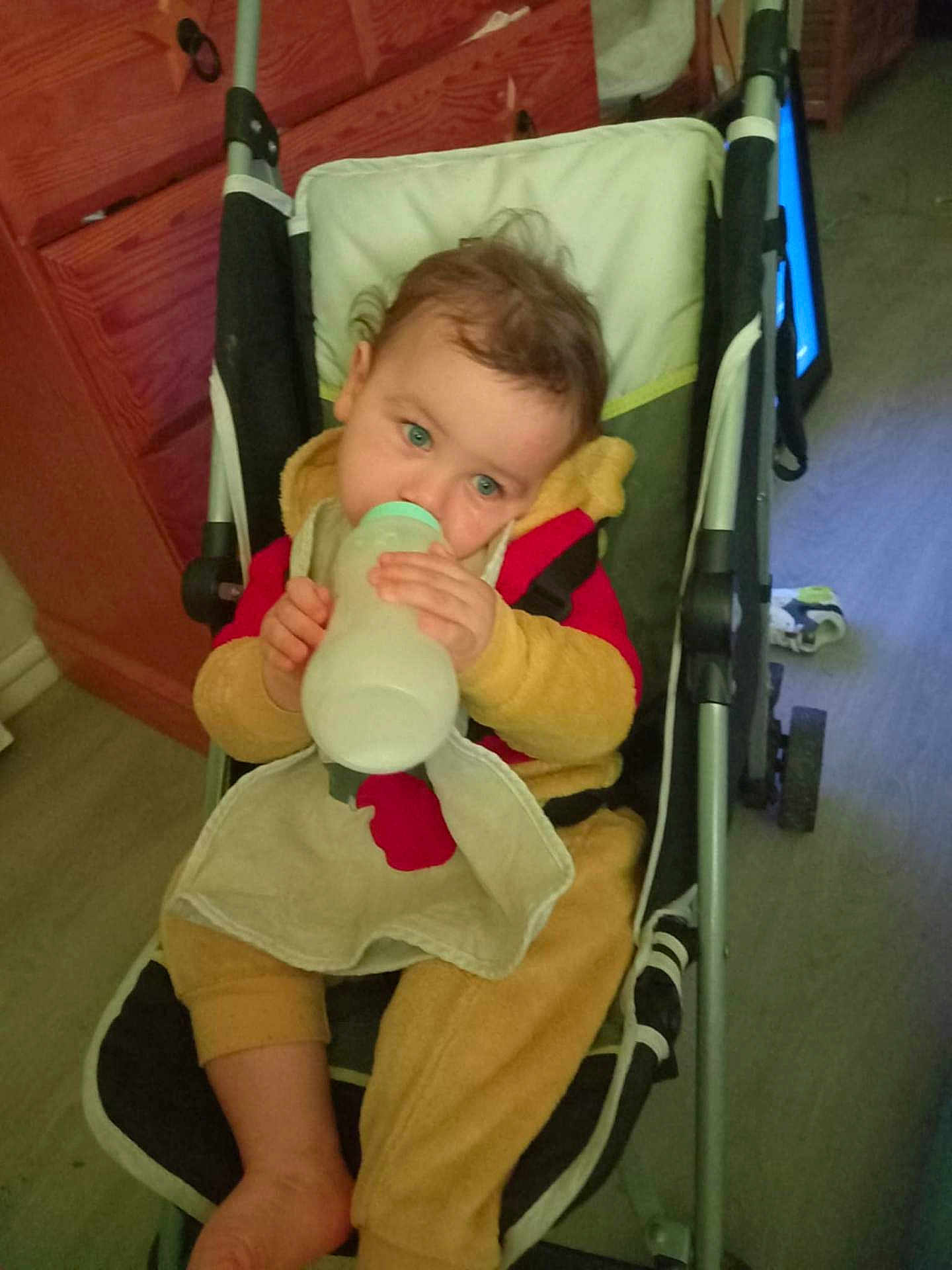 Kenzo participe au concours pour gagner de l'argent avec cette photo : baby, bottle, stroller, child, indoor, drinking, clothing, seat, floor, wooden_cabinet, blanket, foot, person, face, green_eyes, red_clothing, yellow_clothing, baby_bottle, baby_seat, curious