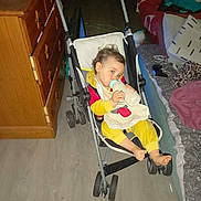 Kenzo participe au concours pour gagner de l'argent avec cette photo : toddler, stroller, bottle, yellow_onesie, child, face, indoor, wooden_dresser, bed, pillow, blanket, floor, footwear, clothing, baby, furniture, person, room, sitting, drinking