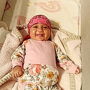 Hazel is registered to the contest to win money with this photo: baby, smiling, pink_bandana, floral_outfit, blanket, lying_down, cute, child, infant, happy, cozy, indoors, soft_texture, face, headwear, person, small_hand, comfort, bed, resting