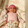 baby, smiling, pink_bandana, floral_outfit, blanket, lying_down, cute, child, infant, happy, cozy, indoors, soft_texture, face, headwear, person, small_hand, comfort, bed, resting