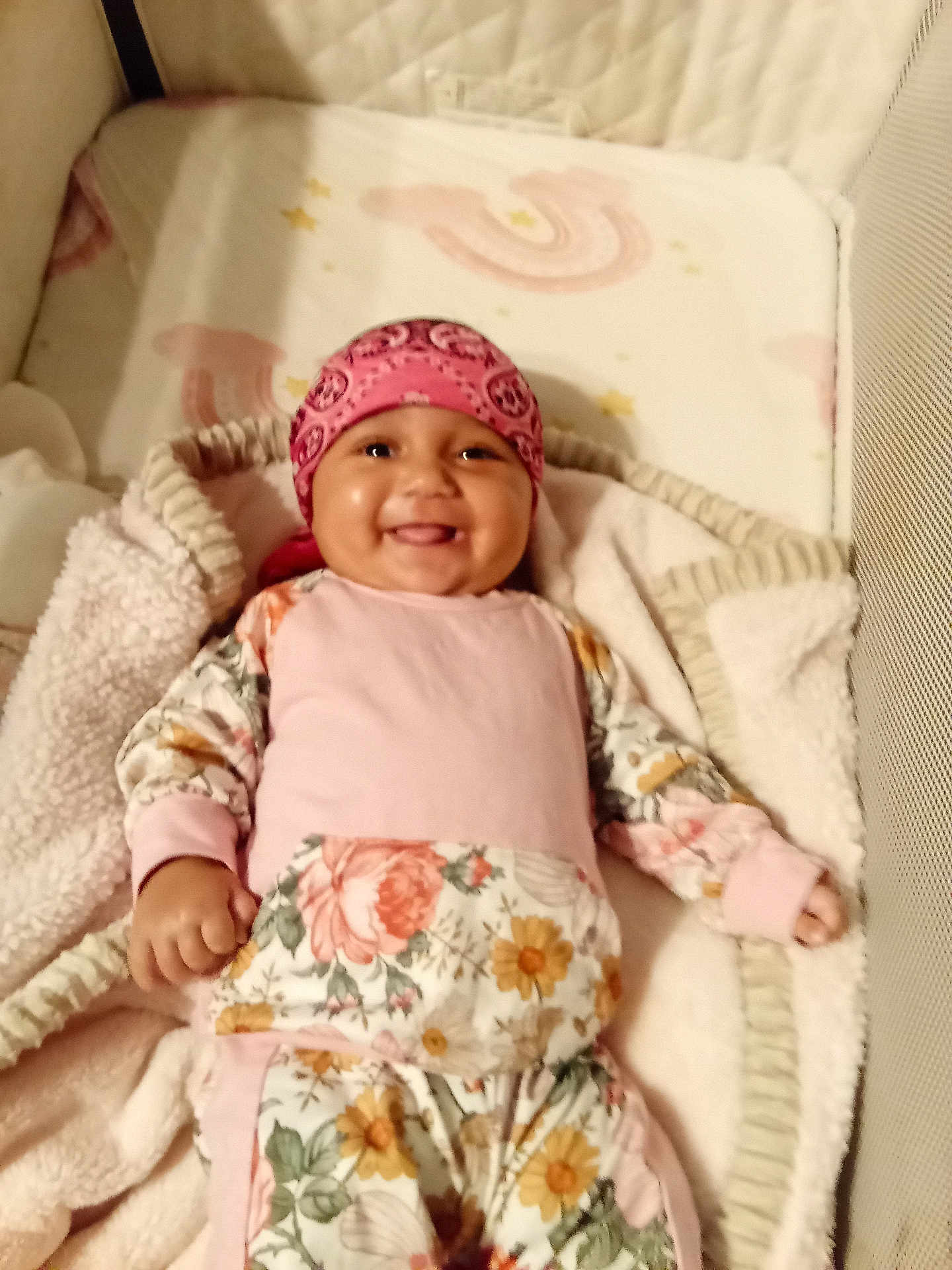 Hazel is registered to the contest to win money with this photo: baby, smiling, pink_bandana, floral_outfit, blanket, lying_down, cute, child, infant, happy, cozy, indoors, soft_texture, face, headwear, person, small_hand, comfort, bed, resting