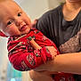 baby, child, person, indoor, holding, pajamas, red_clothing, smile, face, adult, hand, arm, curious, closeup, portrait, young_child, skin, fingernails, blurred_background, casual