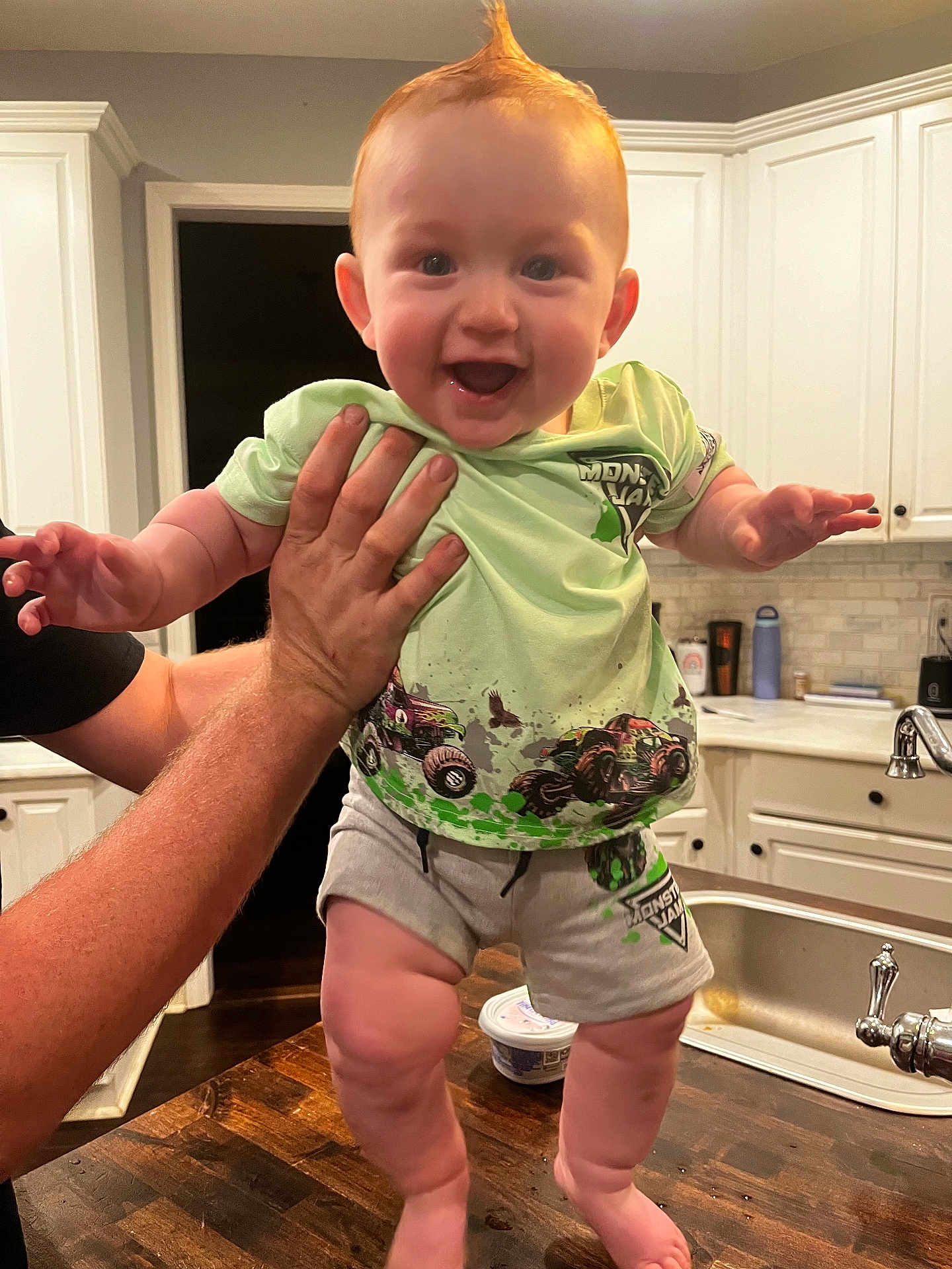 Chase is registered to the contest to win money with this photo: baby, child, smiling, happy, person, kitchen, cabinet, sink, countertop, clothing, shorts, tshirt, hand, table, wood, indoor, facial_expression, hair, holding, playful