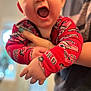 baby, child, infant, red_clothing, hand, face, wide_mouth, surprised_expression, person, adult_hand, holding, indoor, blurred_background, skin, portrait, cute, young, closeup, human, fingernail