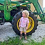 Ryder is registered to the contest to win money with this photo: child, toddler, curly_hair, pink_shirt, cowboy_boots, shorts, tractor, john_deere, large_tire, outdoor, gravel, grass, farm_equipment, yellow, green, side_view, standing, daylight, rural, curious_expression
