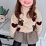 child, girl, smile, portrait, sweater, hearts, bear_motif, skirt, tights, boots, indoor, tile_floor, plant, potted_plant, long_hair, earrings, happy, sitting, cute, candid