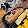 Kirby joined the competition — help win amazing prizes! dog, black_and_white, happy, tongue_out, snack_tray, crackers, cheese, sliced_meat, kitchen, wooden_table, floor, chair, oven, towel, plastic_tray, food, pet, indoor, waiting, curious
