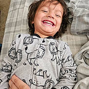 Glenn is registered to the contest to win money with this photo: bed, blanket, child, clothing, cozy, cute, face, hand, happy, indoor, laughing, lion_pattern, pajamas, person, plaid, portrait, sleepwear, smiling, toddler, young