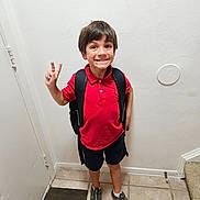 Marcello is registered to the contest to win money with this photo: child, boy, smiling, peace_sign, red_shirt, black_shorts, backpack, sneakers, indoors, tile_floor, door, mat, happy, person, standing, casual_clothing, short_hair, white_wall, home, portrait