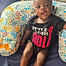 baby, child, infant, portrait, sitting, smiling, indoors, cushion, bed, blanket, black_clothing, graphic_shirt, rock_and_roll, colorful, patterned_fabric, animal_print, face, eyes, legs, photography