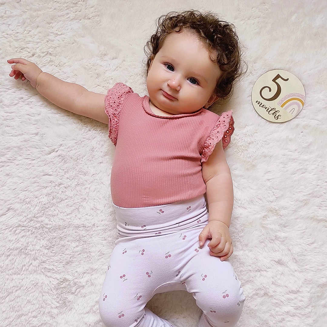 Gemma participe au concours pour gagner de l'argent avec cette photo : 5_months, baby, child, curly_hair, cute, indoors, infant, lying_down, milestone_marker, patterned_pants, person, pink_clothing, portrait, relaxed, skin, smiling, sock, soft_texture, white_rug, young_child