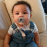 baby, pacifier, denim_overalls, curly_hair, couch, indoor, child, cute, infant, portrait, sitting, clothing, face, hand, eyes, skin, young_child, furniture, relaxed, casual