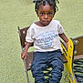 child, toddler, playground, spring_rider, braided_hair, graphic_tee, cargo_pants, sneakers, outdoor, play_equipment, seat, green_surface, expression, looking_at_camera, casual_clothing, young_child, daylight, park, recreation, kid