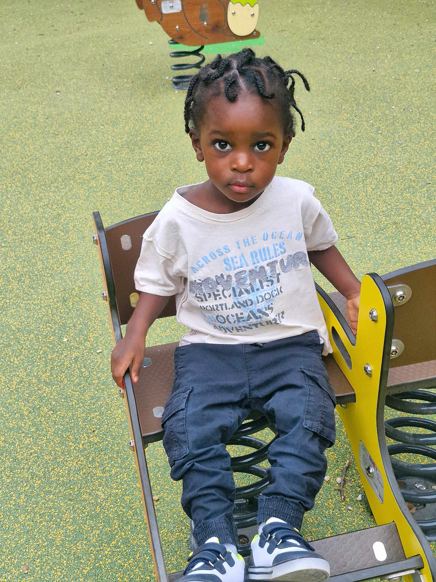 Clayton participe au concours pour gagner de l'argent avec cette photo : child, toddler, playground, spring_rider, braided_hair, graphic_tee, cargo_pants, sneakers, outdoor, play_equipment, seat, green_surface, expression, looking_at_camera, casual_clothing, young_child, daylight, park, recreation, kid