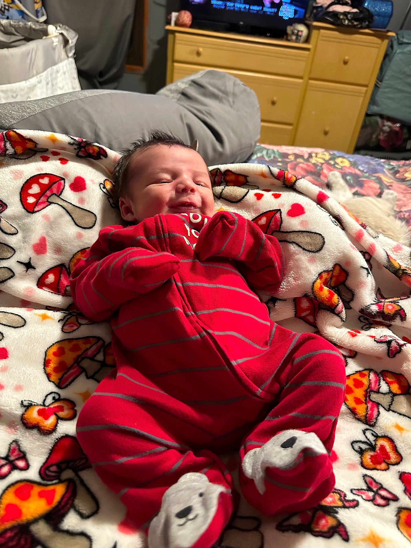 Rimington is registered to the contest to win money with this photo: baby, infant, newborn, blanket, onesie, red_clothing, mushroom_pattern, bed, bedding, pajamas, footed_socks, smiling, face, hair, bedroom, dresser, television, cozy, soft_texture, person