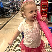 April is registered to the contest to win money with this photo: toddler, child, shopping_cart, pool_noodle, curly_hair, pink_skirt, white_tshirt, shoes, store_aisle, retail_store, skittles_display, floor, smiling, cheeky, indoor, person, toy, grocery_store, shopping, colorful