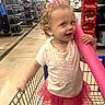 toddler, child, shopping_cart, pool_noodle, curly_hair, pink_skirt, white_tshirt, shoes, store_aisle, retail_store, skittles_display, floor, smiling, cheeky, indoor, person, toy, grocery_store, shopping, colorful