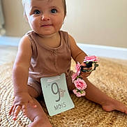 Melia a rejoint le concours — aidez-le/la à gagner de superbes lots ! baby, child, infant, sitting, milestone_card, pink_flowers, brown_outfit, woven_rug, cute, portrait, indoor, smiling, person, hand, foot, head, face, blue_eyes, decor, soft_lighting