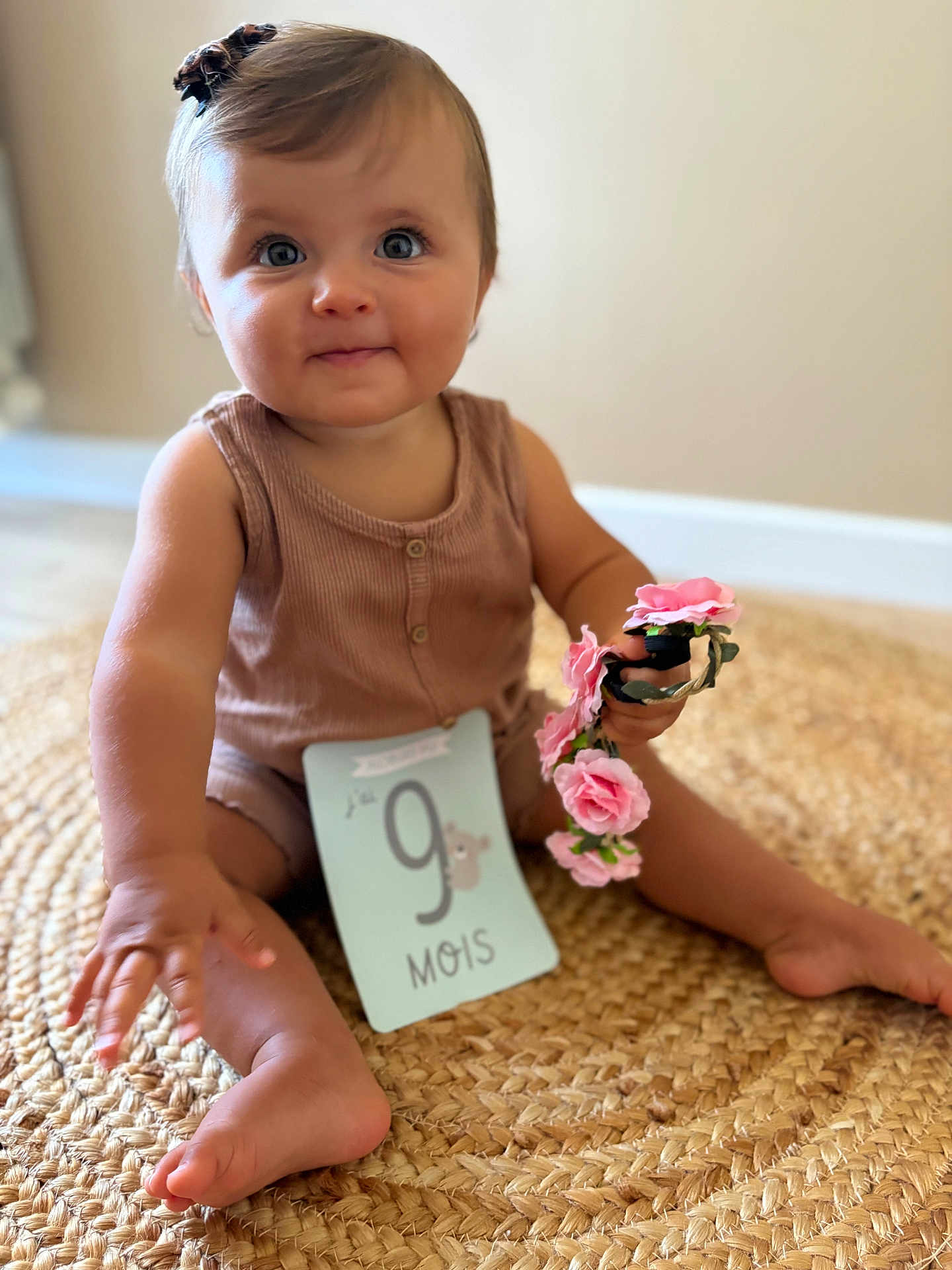 Melia a rejoint le concours — aidez-le/la à gagner de superbes lots ! baby, child, infant, sitting, milestone_card, pink_flowers, brown_outfit, woven_rug, cute, portrait, indoor, smiling, person, hand, foot, head, face, blue_eyes, decor, soft_lighting
