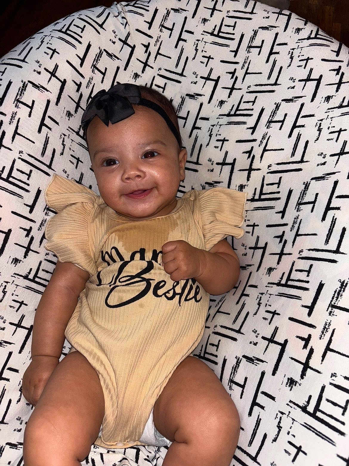 Avani is registered to the contest to win money with this photo: baby, infant, smiling, face, headband, clothing, onesie, patterned_background, cushion, person, cute, happy, skin, fist, sitting, indoors, child, young, comfortable, portrait