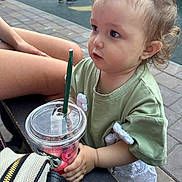 Erika is registered to the contest to win money with this photo: toddler, child, starbucks, cup, snack, green_tshirt, curly_hair, outdoor, bench, legs, pavement, person, hand, plastic_lid, straw, daylight, casual_clothing, park, young_child, curious