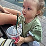 toddler, child, starbucks, cup, snack, green_tshirt, curly_hair, outdoor, bench, legs, pavement, person, hand, plastic_lid, straw, daylight, casual_clothing, park, young_child, curious
