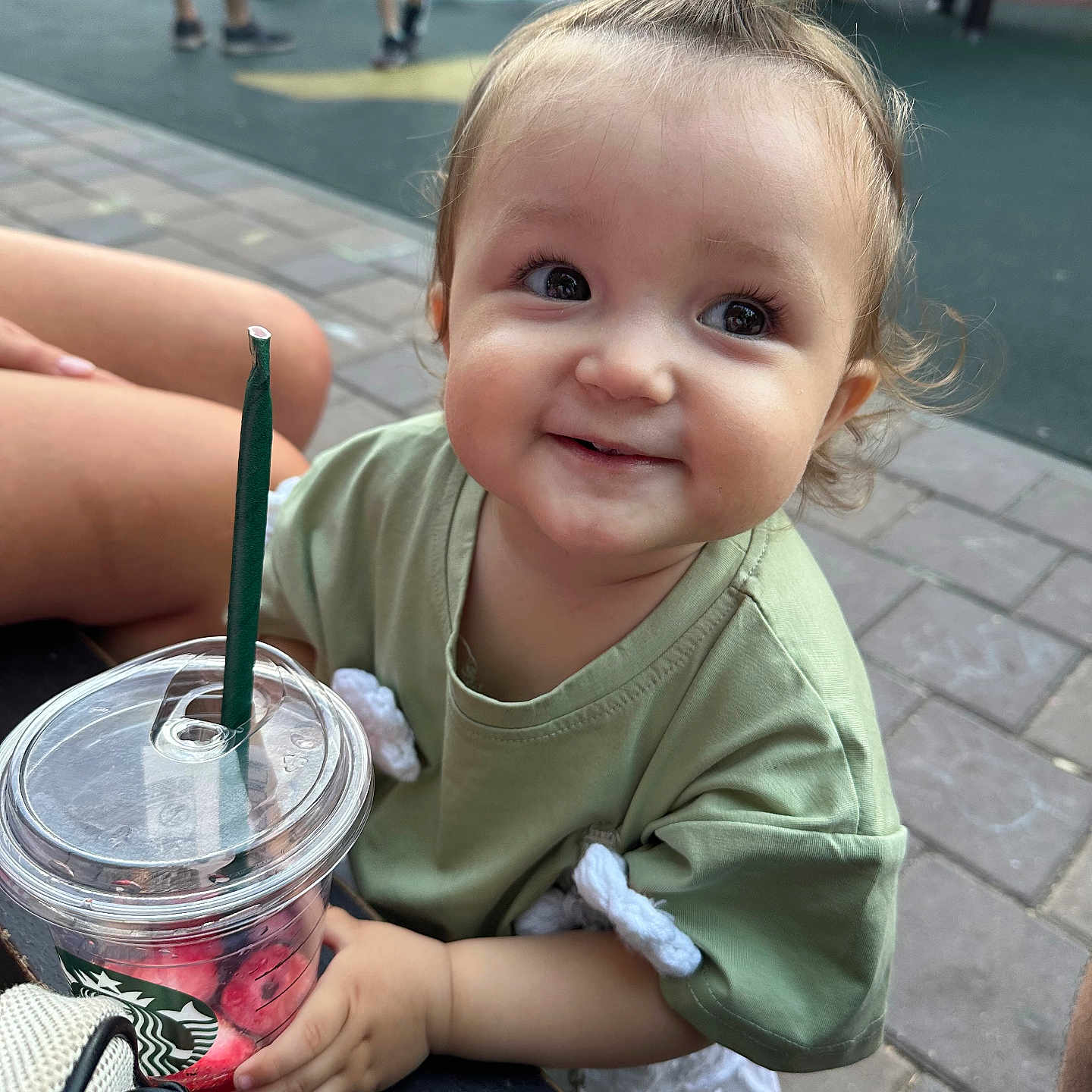 Erika is registered to the contest to win money with this photo: bench, casual_clothing, child, cute, daylight, drink, green_shirt, hair_tie, hand, happy, outdoor, pavement, person, playground, sitting, smile, starbucks_cup, street, toddler, young_child