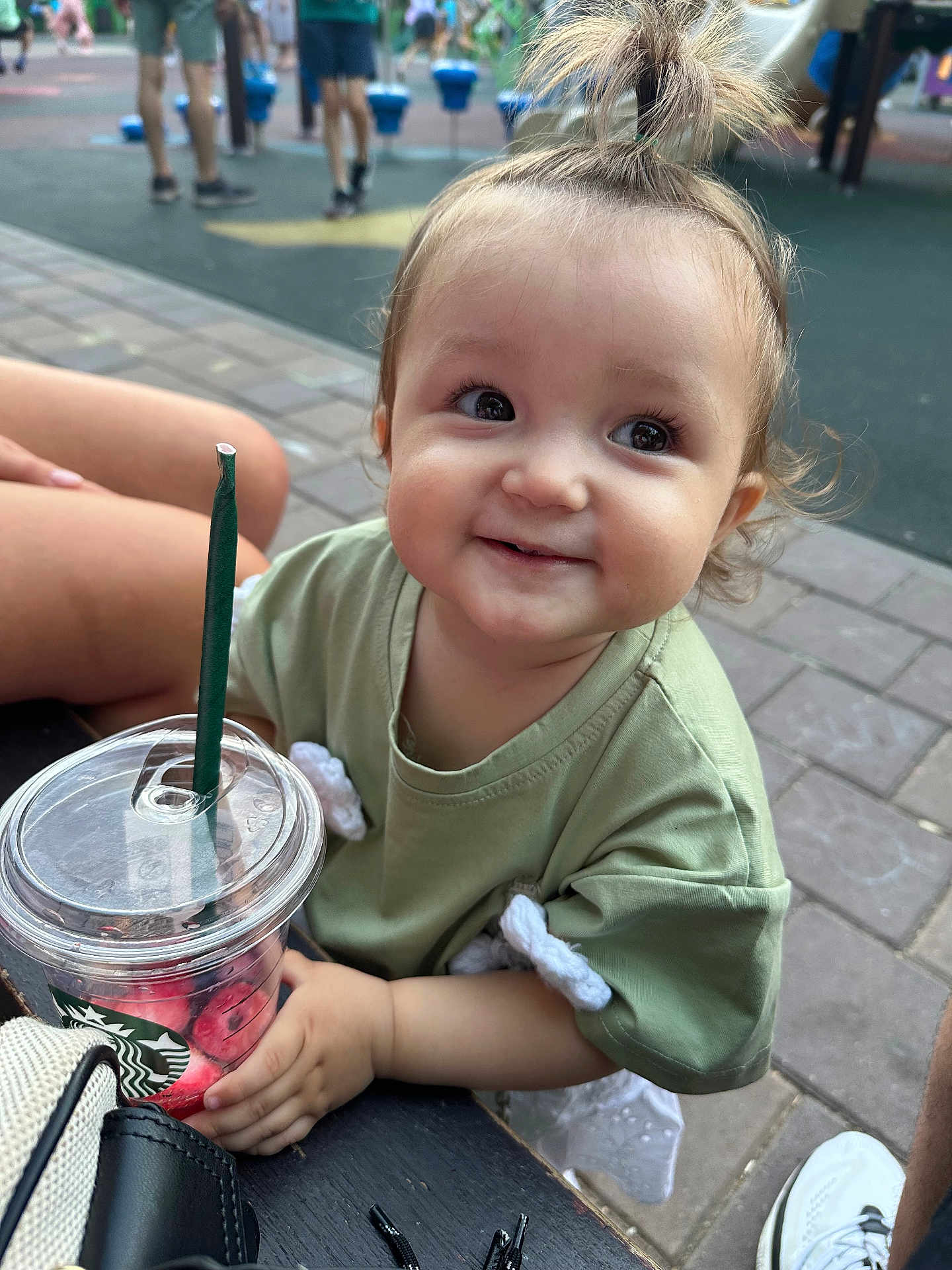 Erika is registered to the contest to win money with this photo: toddler, child, smile, green_shirt, starbucks_cup, drink, outdoor, bench, playground, pavement, person, hair_tie, happy, cute, daylight, casual_clothing, sitting, hand, young_child, street