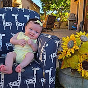 Remi is registered to the contest to win money with this photo: baby, child, yellow_outfit, giraffe_pattern, armchair, outdoor, grass, sunflowers, flower_pot, smiling, headband, furniture, tree, sunlight, garden, patio, blue_sky, happy, person, cute