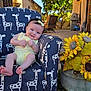 baby, child, yellow_outfit, giraffe_pattern, armchair, outdoor, grass, sunflowers, flower_pot, smiling, headband, furniture, tree, sunlight, garden, patio, blue_sky, happy, person, cute