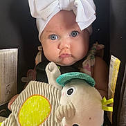 Violet is registered to the contest to win money with this photo: baby, child, car_seat, white_bow, plush_toy, blue_eyes, face, clothing, toddler, infant, cute, portrait, soft_toy, seatbelt, indoor, person, headwear, expression, closeup, seat