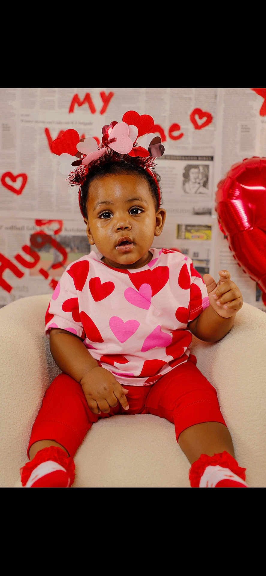 Zanari is registered to the contest to win money with this photo: accessory, baby, balloon, celebration, chair, child, cute, expression, headband, hearts, holiday_theme, newspaper_backdrop, outfit, patterned_shirt, pink_and_red, portrait, sitting, studio_background, toddler, valentine