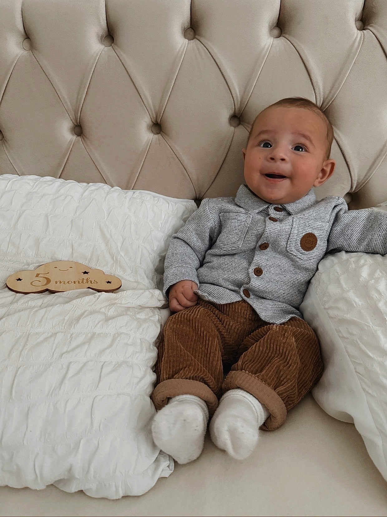 Ibrahim participe au concours pour gagner de l'argent avec cette photo : baby, infant, smiling, couch, pillow, clothing, corduroy, shirt, button_up, milestone, wooden_sign, face, happy, sitting, indoor, soft_lighting, head, feet, white_socks, tufted_couch