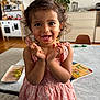 toddler, child, smiling, happy, curly_hair, dress, indoor, living_room, table, toys, book, grapes, person, furniture, plant, chair, face, hands, floor, home
