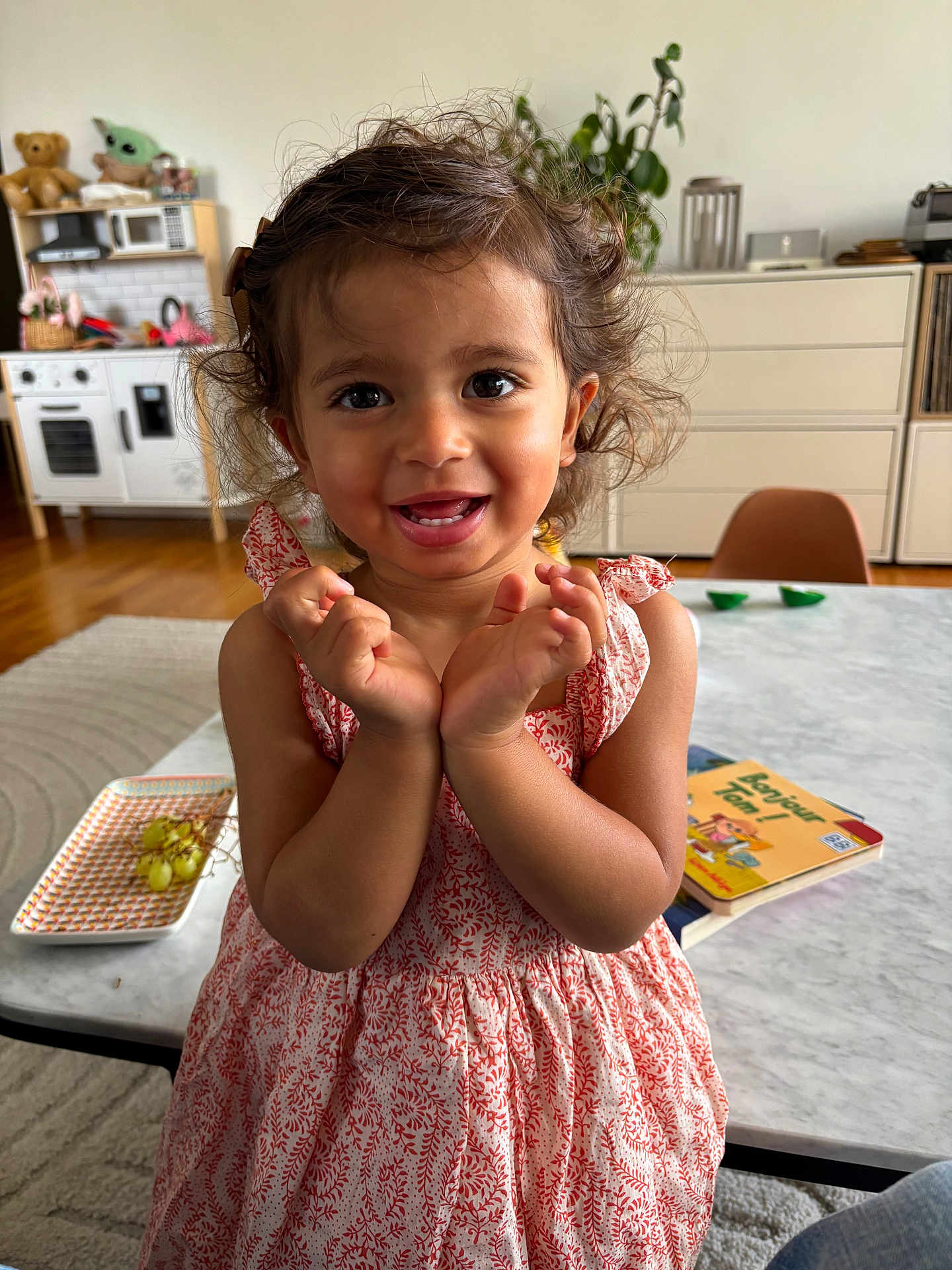 Ezzah participe au concours pour gagner de l'argent avec cette photo : toddler, child, smiling, happy, curly_hair, dress, indoor, living_room, table, toys, book, grapes, person, furniture, plant, chair, face, hands, floor, home