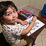 art_supplies, beige_pants, black_hair, chair, child, colorful, crayons, door, drawing, expression, floor, home, indoor, looking_up, patterned_shirt, seated, short_sleeve_shirt, table, tile_floor, young_child