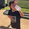 bench, black_tshirt, blue_shoes, boy, casual_clothing, child, daytime, drinking, grass, outdoor, park, pavement, plaid_shorts, playground, portrait, shadow, sunny, trash_can, water_bottle, young_child