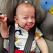 Antonio participe au concours pour gagner de l'argent avec cette photo : baby, car_seat, child, face, tongue_out, playful, smiling, clothing, seatbelt, infant, red_pants, white_shirt, patterned_shirt, toy, colorful, expression, cute, indoor, closeup, cheeky