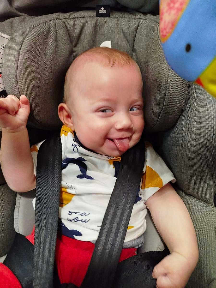Antonio participe au concours pour gagner de l'argent avec cette photo : baby, car_seat, child, face, tongue_out, playful, smiling, clothing, seatbelt, infant, red_pants, white_shirt, patterned_shirt, toy, colorful, expression, cute, indoor, closeup, cheeky