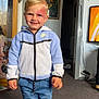 child, boy, smiling, indoor, nike_jacket, blue_jeans, carpet, cabinet, framed_picture, television, home, person, socks, standing, birthmark, doorway, curtains, furniture, room, casual_clothing