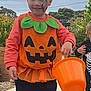 boots, boy, brick_wall, child, cloudy_sky, costume, face_paint, fall, green_leaves, halloween, holiday, orange_bucket, outdoor, playful, pumpkin_costume, sidewalk, skeleton_costume, smiling, trees, trick_or_treat