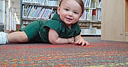Cannon is registered to the contest to win money with this photo: toddler, child, smile, library, bookshelf, carpet, indoors, green_clothing, white_shoes, floor, person, young_child, cute, happy, playful, reading_area, furniture, window, daylight, sitting