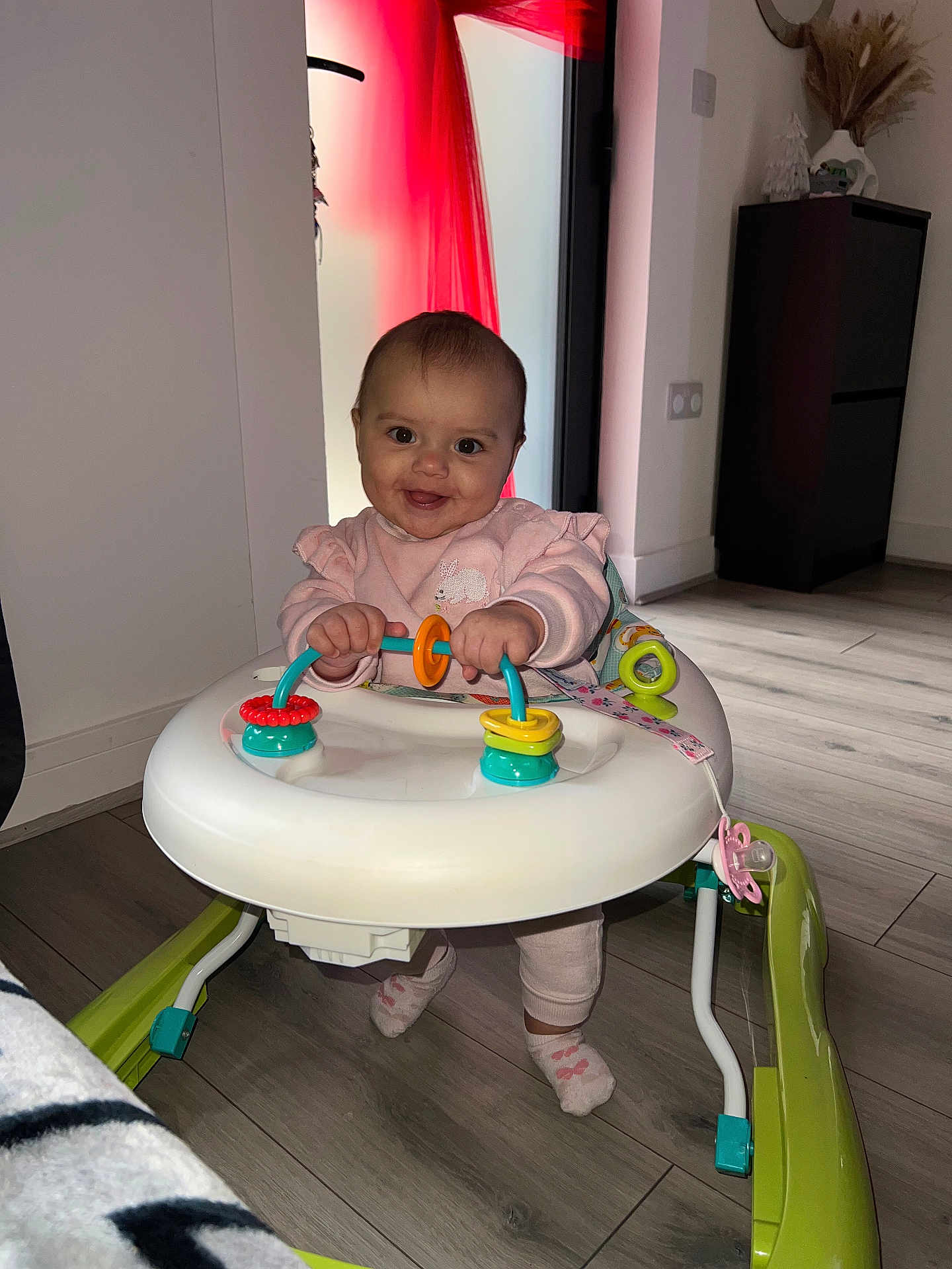Maddison joined the competition — help win amazing prizes! baby, child, walker, toy, indoor, floor, pink_clothing, smiling, happy, cute, infant, person, play, furniture, home, wooden_floor, socks, pacifier, holding, face