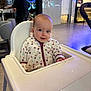 baby, high_chair, child, indoor, person, jacket, heart_pattern, face, expression, furniture, table, stroller, blurred_background, modern_interior, happy, cute, seated, curious, cozy, casual