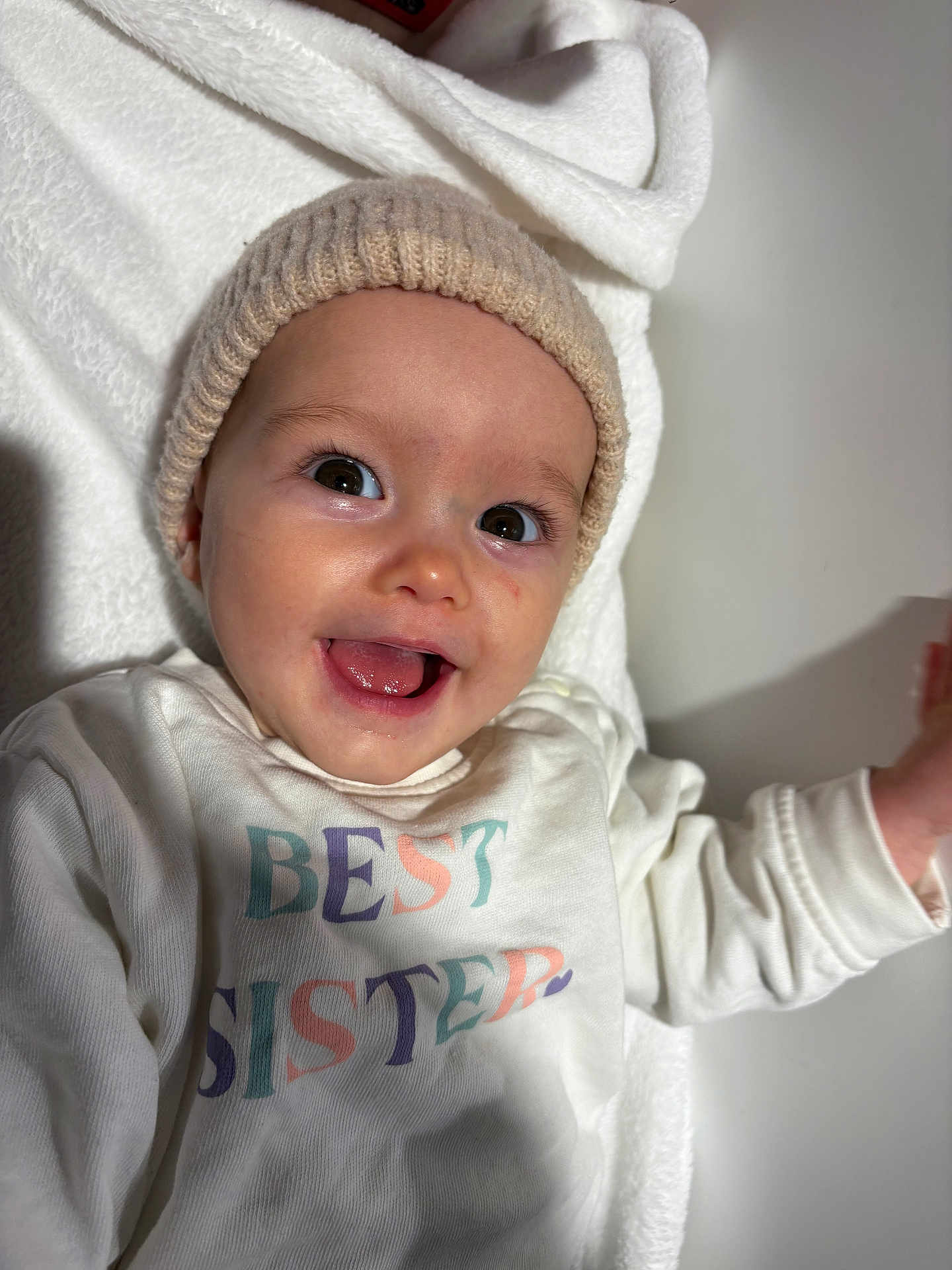 Mylie participe au concours pour gagner de l'argent avec cette photo : baby, child, infant, smiling, hat, knit_hat, white_shirt, text_on_clothing, best_sister, tongue_out, happy, portrait, face, eyes, blanket, soft_texture, indoors, close_up, person, cute