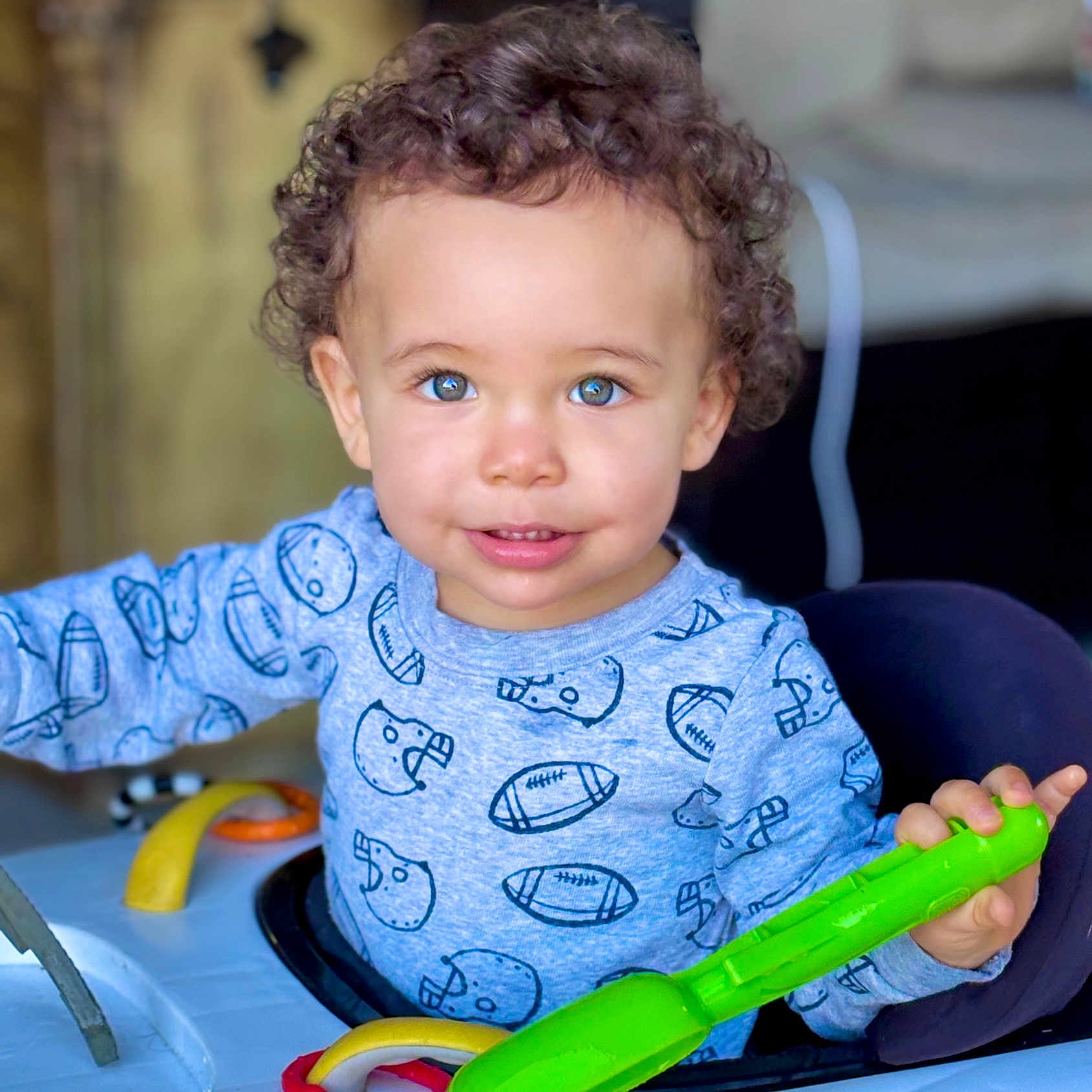 Madden is registered to the contest to win money with this photo: toddler, child, curly_hair, blue_eyes, smiling, playing, toy, green_toy, blue_shirt, football_print, indoor, baby_walker, happy, cute, person, face, hand, clothing, young_child, portrait