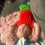 baby, sleeping, pillow, red_shirt, green_pants, blanket, couch, indoor, child, peaceful, resting, soft_texture, cozy, infant, relaxed, home, furniture, person, toddler, nap