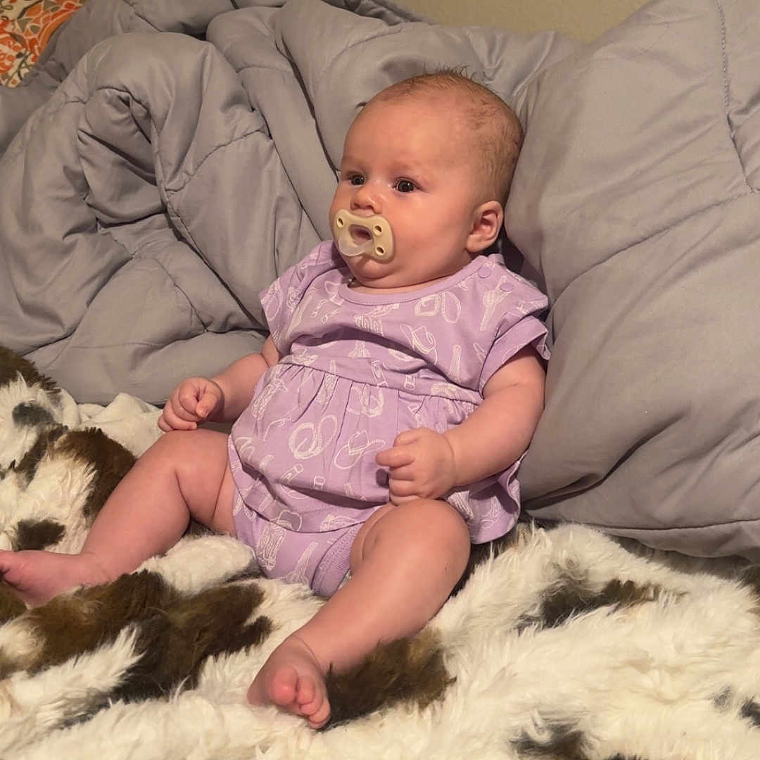 Daisy is registered to the contest to win money with this photo: baby, infant, pacifier, purple_clothing, blanket, fur_blanket, quilt, bedroom, wall, poster, cute, sitting, child, indoors, comfort, soft, young, person, relax, cozy